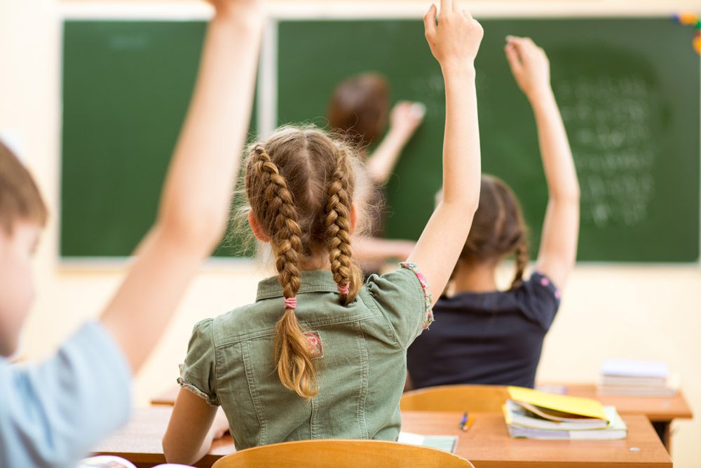 A group of children raising their hands in front of a chalkboard.